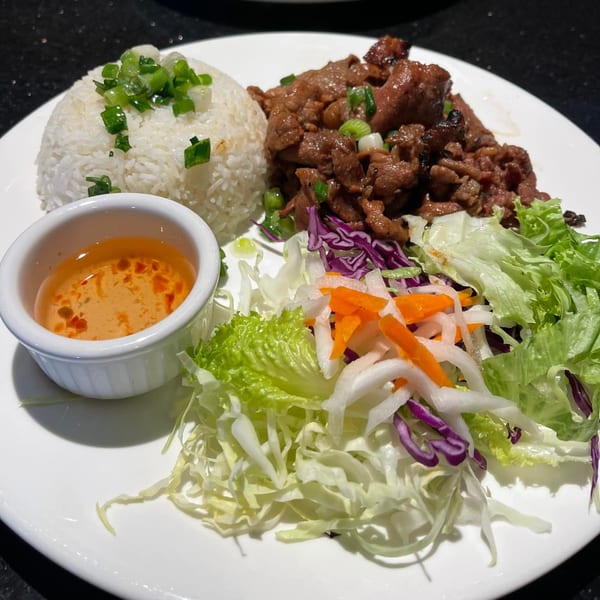 Vietnamese grilled pork with rice, fresh salad, and a side of dipping sauce on a white plate.