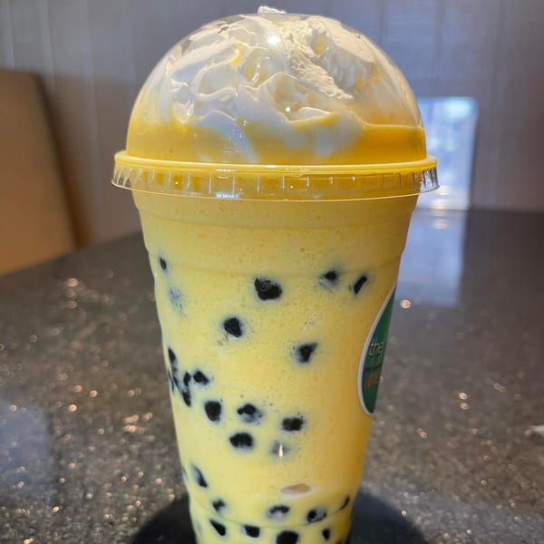 A yellow mango bubble tea with whipped cream and black boba pearls in a clear plastic cup.