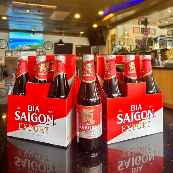 Three six-packs of Bia Saigon Export beer bottles on a reflective counter at The Phở Shop.