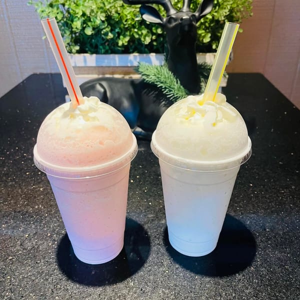 Two Vietnamese smoothies, one pink and one white, topped with whipped cream and straws on a dark counter.