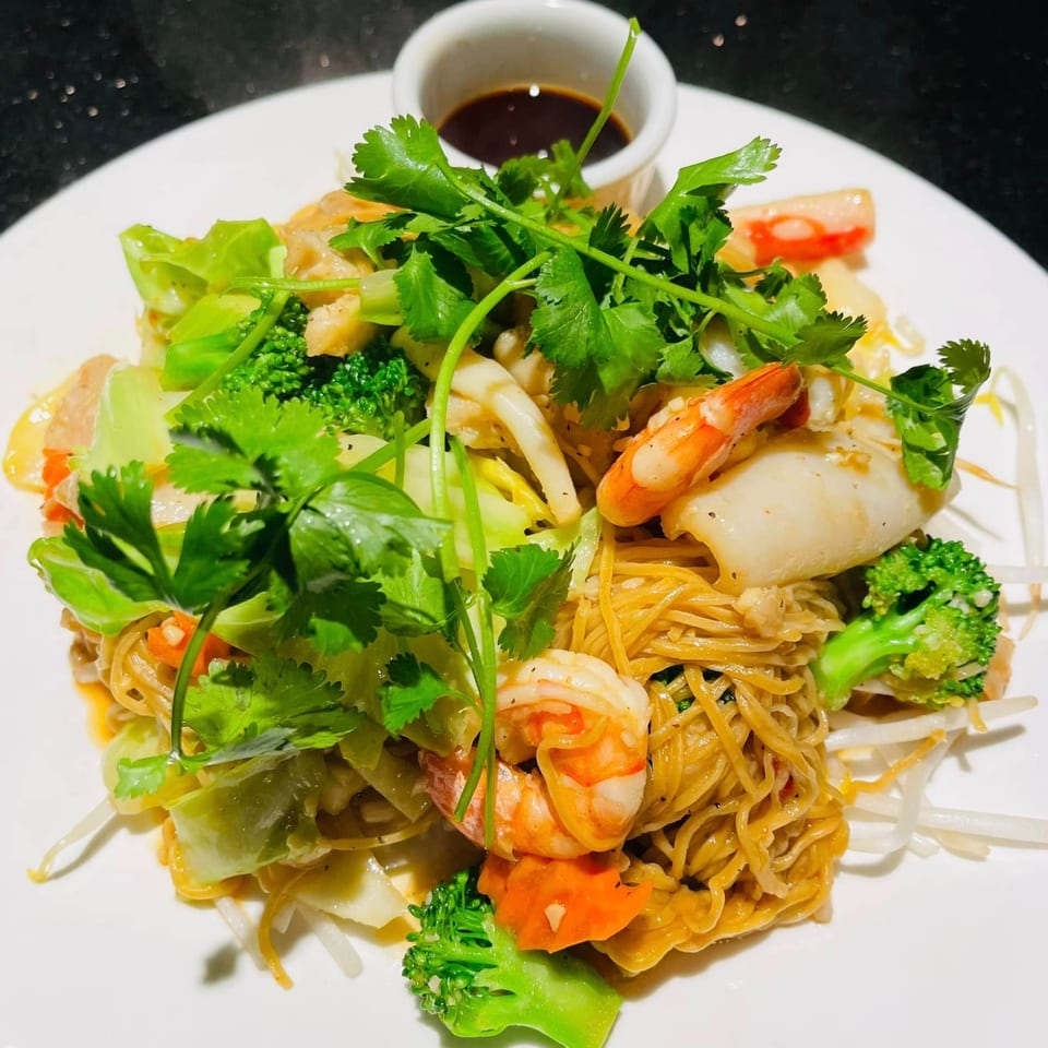 Crispy egg noodles with shrimp, squid, broccoli, and cilantro, served with a side of dark sauce.
